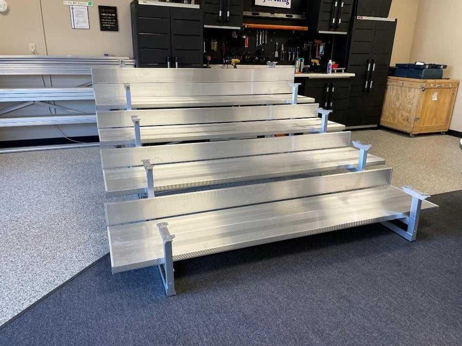 A fully assembled four-row aluminum stationary bleacher standing on a dealer showroom floor with no guardrail, seat planks carried on cast plank clips.