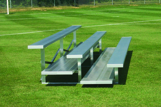 A three-row aluminum bleacher with a seat plank and separate foot plank on each row, standing free on mown grass at the edge of a sports field.
