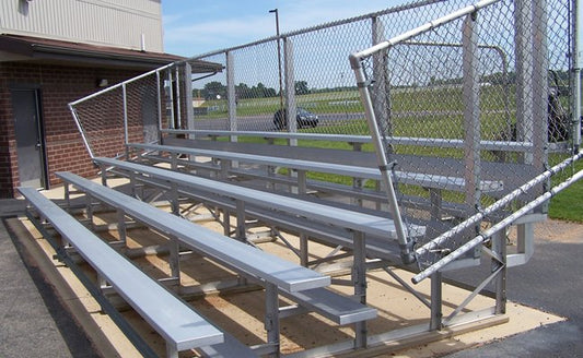 A five-row aluminum bleacher with chain-link back and end guardrails on a concrete pad at the corner of a brick school building, an access gate open at the right end.