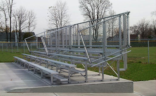 A five-row aluminum bleacher with chain-link back and end guardrails raised on a concrete pad at a ball field on an overcast day; trim level is not determinable from the photo.