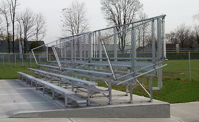A five-row aluminum bleacher with chain-link back and end guardrails raised on a concrete pad at a ball field on an overcast day; trim level is not determinable from the photo.