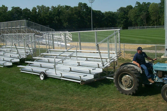 A five-row transportable bleacher on a wheeled towing frame being pulled across a ball field by a tractor, with chain-link guardrails and a second identical unit parked behind.