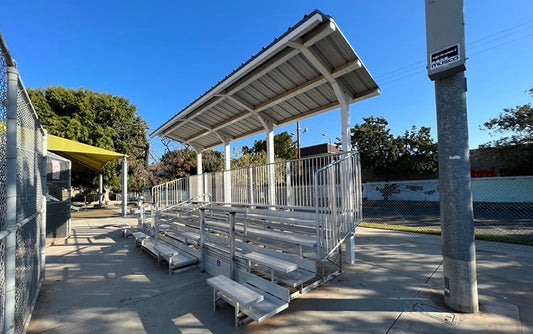 A five-row aluminum bleacher under a fixed metal shade canopy, with vertical picket guardrails, end handrails and a blue ADA space marking on the front deck, on a school hardcourt; the canopy is a configuration no listed product includes.