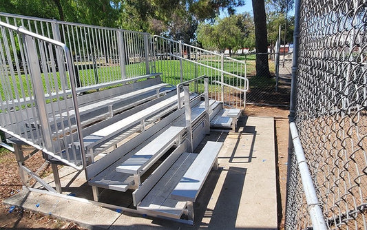 Five-row aluminum bleacher with vertical picket guardrail, double foot planks on every row and an end handrail, on a concrete pad beside a grass park field; no ADA placard visible in frame.
