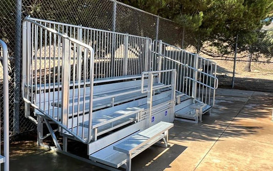 Five-row bleacher with vertical picket guardrail carrying a blue ADA wheelchair placard on the front row, a separate ADA companion bench at pad level and an aisle handrail, on concrete under pines.