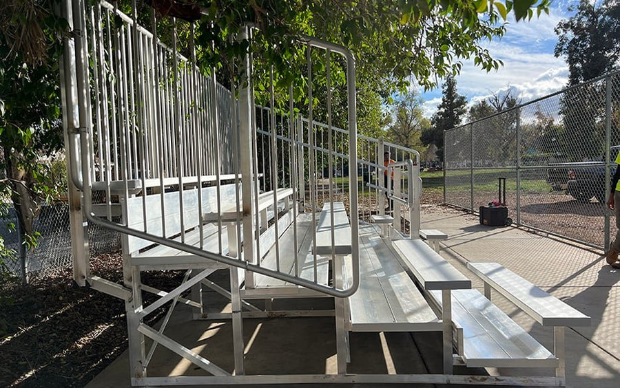 Five-row bleacher with vertical picket guardrail and an aisle handrail on a concrete pad under trees, chain link site fencing and a park path behind; a bystander's legs are at the right edge.