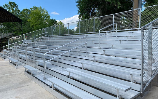 Tall aluminum bleacher bank with a chain link guardrail across the back and both ends, an aisle handrail and foot planks on every row, on a concrete pad at a ballfield; too oblique to count rows reliably, roughly eight to ten.