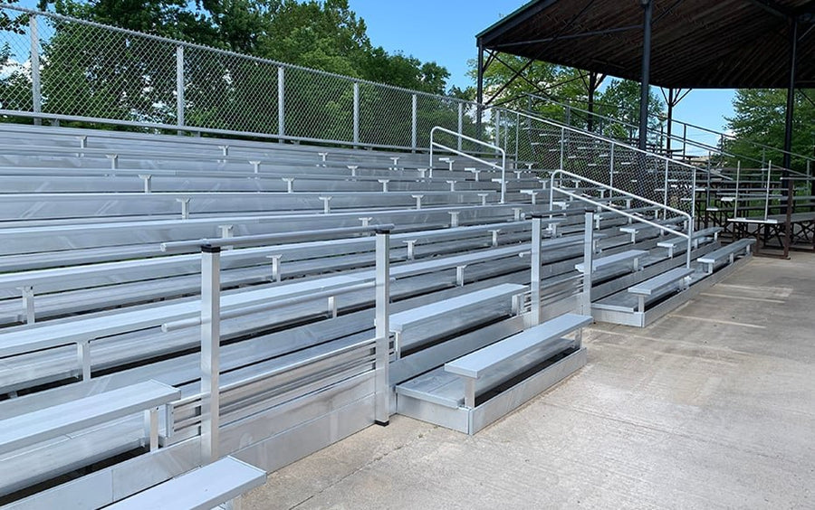 Front-left view of the same tall chain link guardrail bleacher bank with its aisle handrail, a roofed grandstand at the right edge; rows are visible but not countable with confidence.
