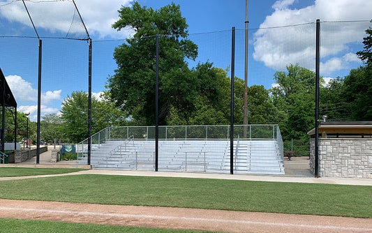 The bleacher bank seen from the infield through backstop netting, chain link guardrail along the top, with a stone dugout building at the right.
