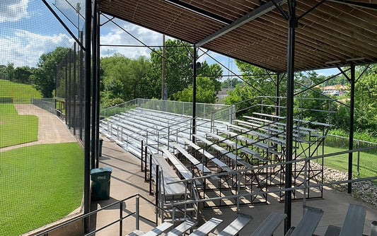 From under the roofed grandstand looking across many rows of aluminum bleachers with a chain link guardrail behind, wood roof deck overhead and the field at left.