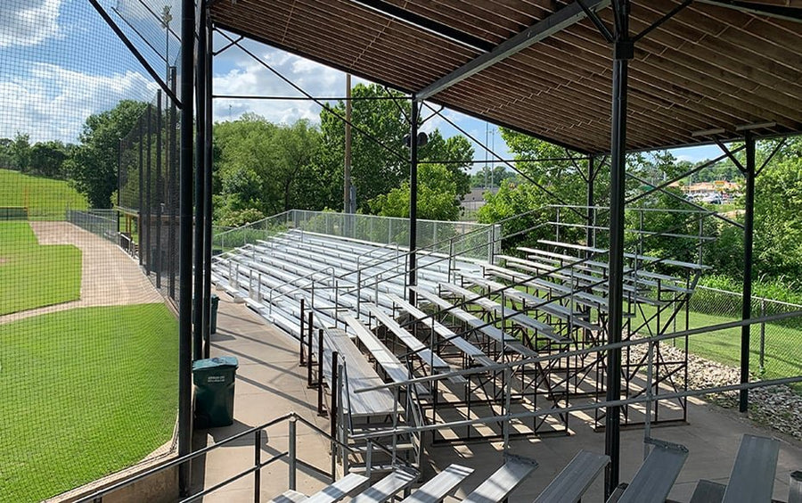 From under the roofed grandstand looking across many rows of aluminum bleachers with a chain link guardrail behind, wood roof deck overhead and the field at left.