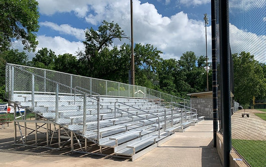 Three-quarter view of the tall bleacher bank showing the stepped aluminum end frame and understructure, chain link guardrail on the back and end, and an aisle handrail up the middle.