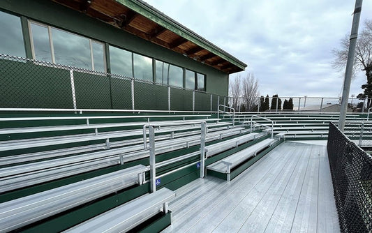 Low-rise aluminum bleachers with dark green riser panels and full aluminum decking on a wood deck beside a green ballpark press building, pipe aisle handrails and blue wheelchair-space decals on the front row.