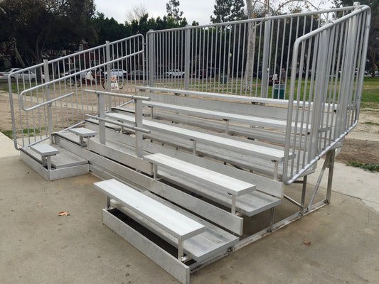 Freestanding five-row aluminum bleacher with vertical picket guardrail across the rear and both ends, seat planks over separate footboards, a center aisle with steps and a side entry landing on a park concrete pad.
