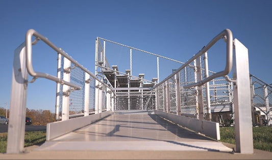 Head-on view straight up an aluminum accessible ramp with pipe handrails leading to a bleacher fitted with chain link guardrail panels.