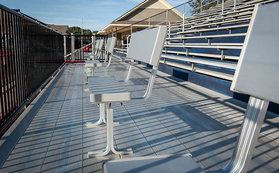 Pedestal-mounted aluminum bench seats with backrests lined along a decked platform in front of bleacher rows, black picket railing behind them.
