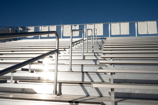 Backlit close-up of aluminum bleacher seat planks and footboards with pipe aisle handrails, chain link guardrail with blue windscreen panels along the top row.