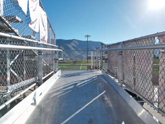 Looking along an aluminum accessible ramp deck with chain link infill guardrail panels on both sides, out toward the field and mountains.