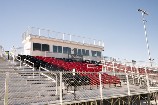Press box with sliding windows and a railed rooftop camera deck sitting atop a grandstand, red and black chair-back seating below and a chain link guardrail across the front.